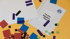 Tactile Algebra Tiles magnetic board with a variety of tiles scattered around, all on a yellow background.