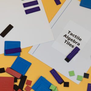 Tactile Algebra Tiles magnetic board with a variety of tiles scattered around, all on a yellow background.