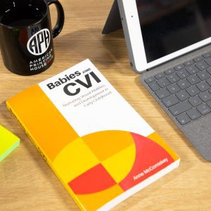 Babies with CVI book sits atop a desk, next to a laptop, APH mug, and pad of paper and pen