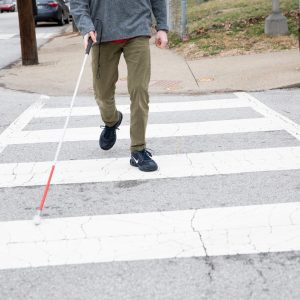 Photo of the legs and feet of a person with a white cane crossing at a cross walk.