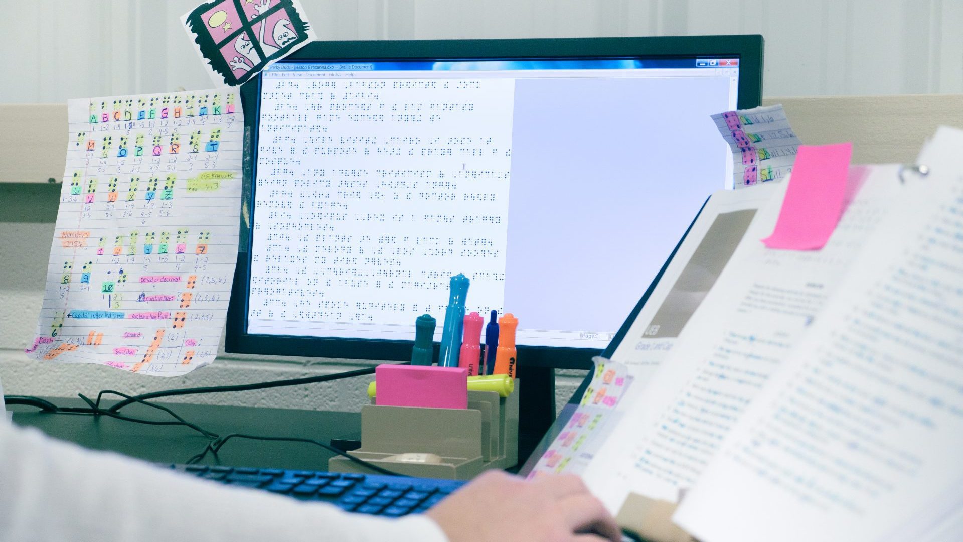 A work station with a computer displaying braille, a desk organizer holding highlighters and Post-It notes, a paper taped to the monitor with color coded notes on the braille alphabet, and a little printed illustrations of funny ghosts. A hand is reaching out to turn the page of a marked up text in a binder