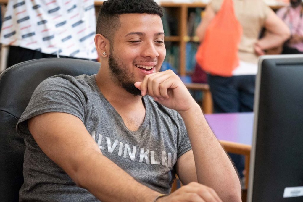 An older teen boy sits smiling at a computer in a classroom.