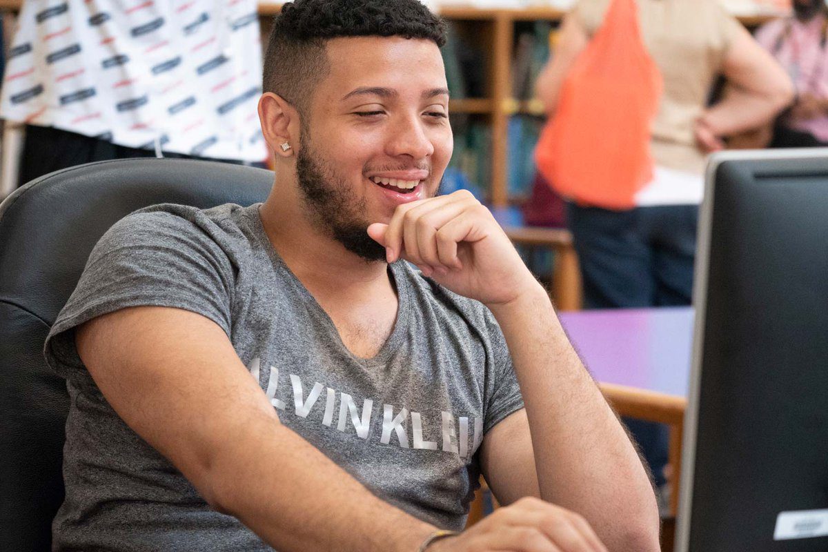 An older teen boy sits smiling at a computer in a classroom.