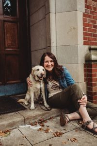 A smiling woman sits on a step with her arm around her guide dog, a yellow lab.