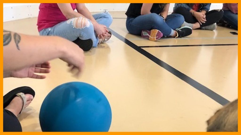 Adults sitting on a gym floor rolling a goalball.