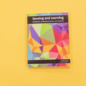 Sensing and Learning Guidebook, Assessment Forms, and Routines resting on a yellow background. The cover of the book showcases variations of pomegranate, teal, gold, and green.