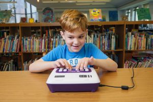 A boy with an excited expression places his hands on Polly, an electronic braille learning device.