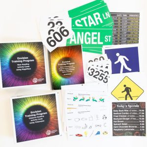 Envision Kit without Optical Aids opened to show training curriculum, activity cards, signage, restaurant menu, and additional large print resources.