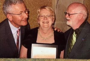 A smiling woman with short hair holds a plaque with two smiling men, who are looking at each other, stand beside her. 
