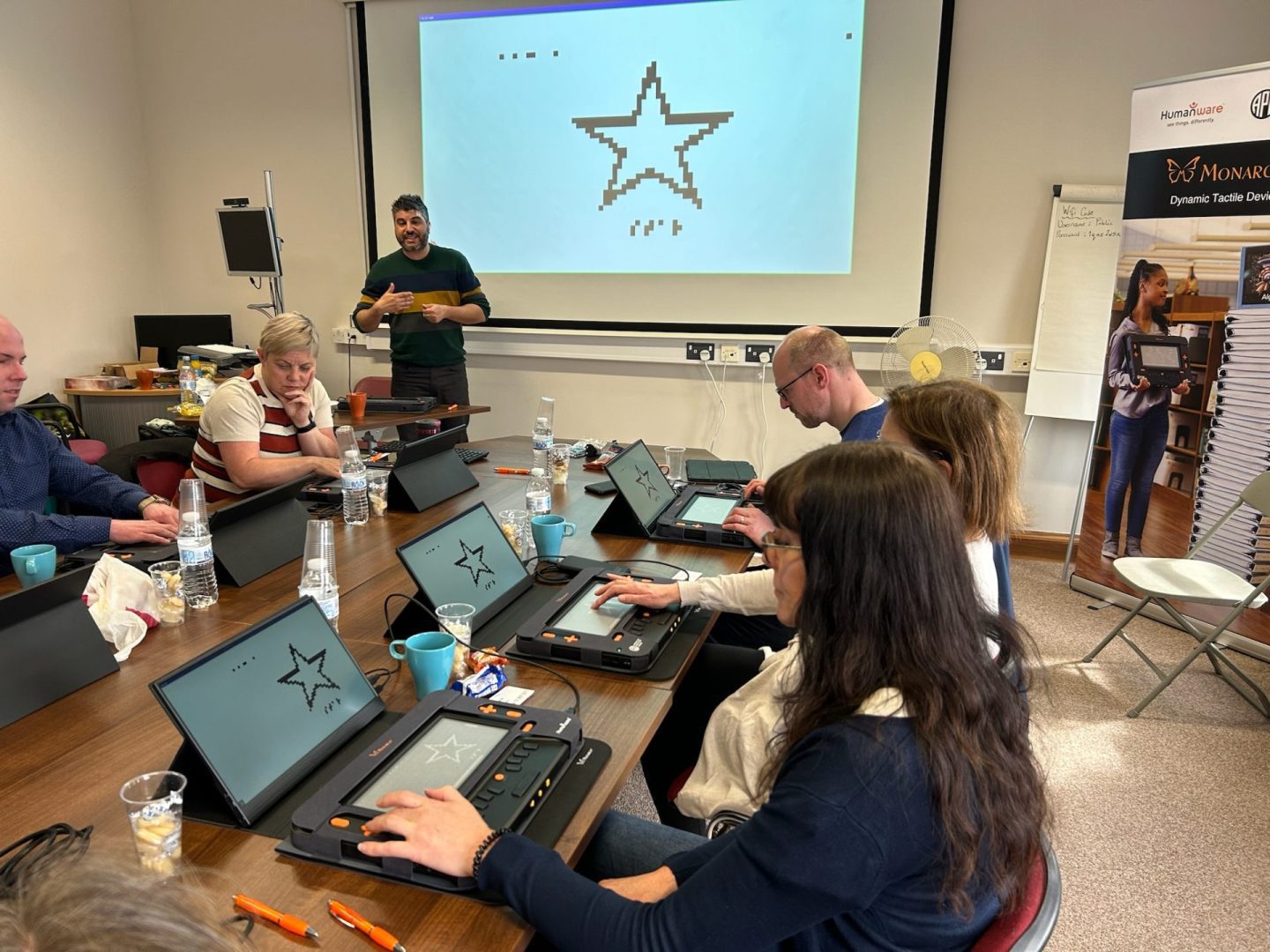 A man speaks to a group seated at a conference room table, each with a Monarch multiline refreshable braille display and a small monitor in front of them. Behind the man is a large screen displaying a star, which is also appearing on the small monitors and as a tactile graphic on the Monarch.