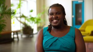Denna Lambert smiles in a well-lit room where plants and a yellow sofa are visible behind her.