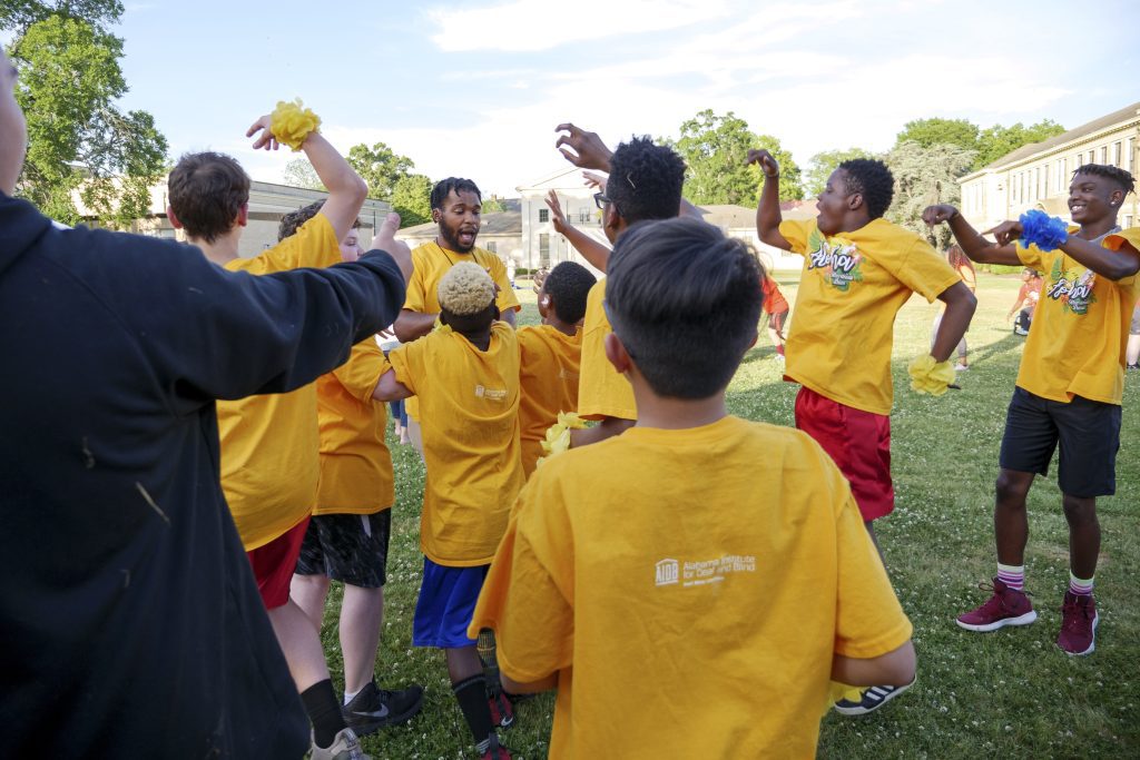 A group of students in yellow shirts jump and cheer with a teacher as they stand in the grass.
