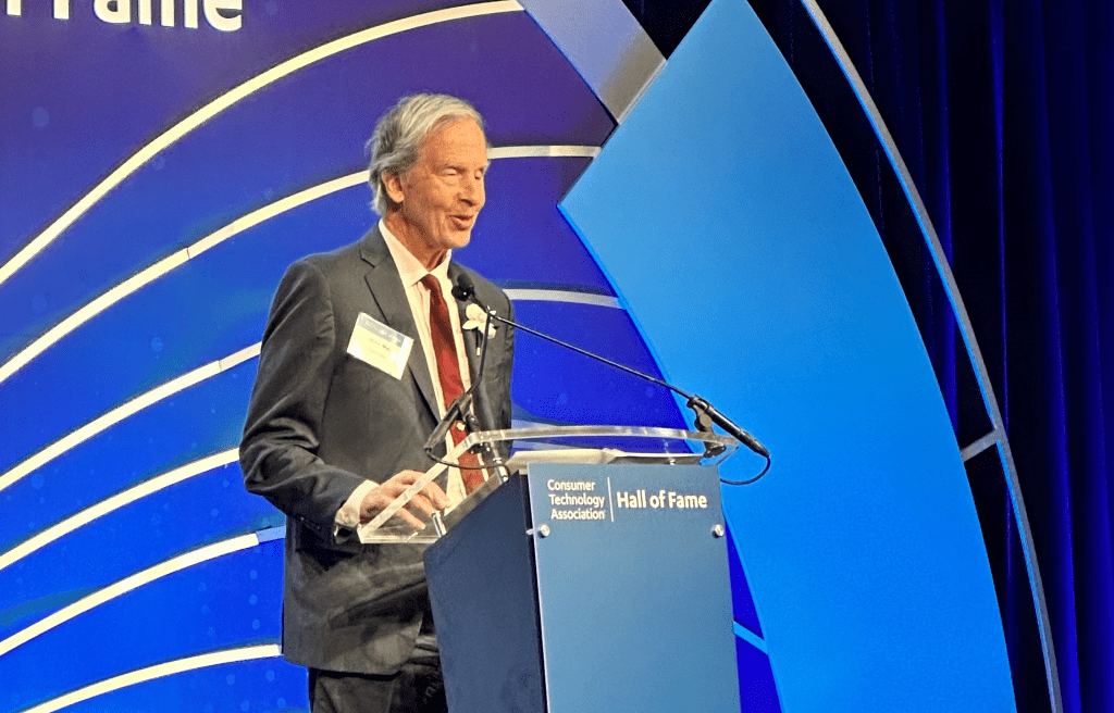 Mike May wears a suit as he speaks from a podium with the words
