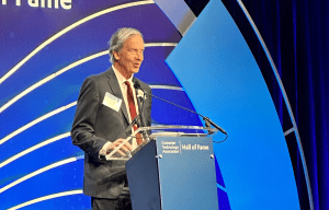 Mike May wears a suit as he speaks from a podium with the words 