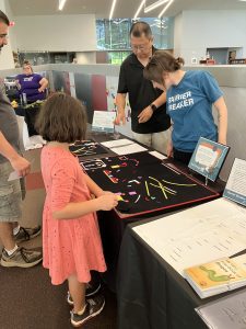 A young girl standing near a man plays with pieces of the Picture Maker Wheatley Tactile Diagramming Kit, which are resting on a table, while two APH volunteers speak to her and the man from behind the table.