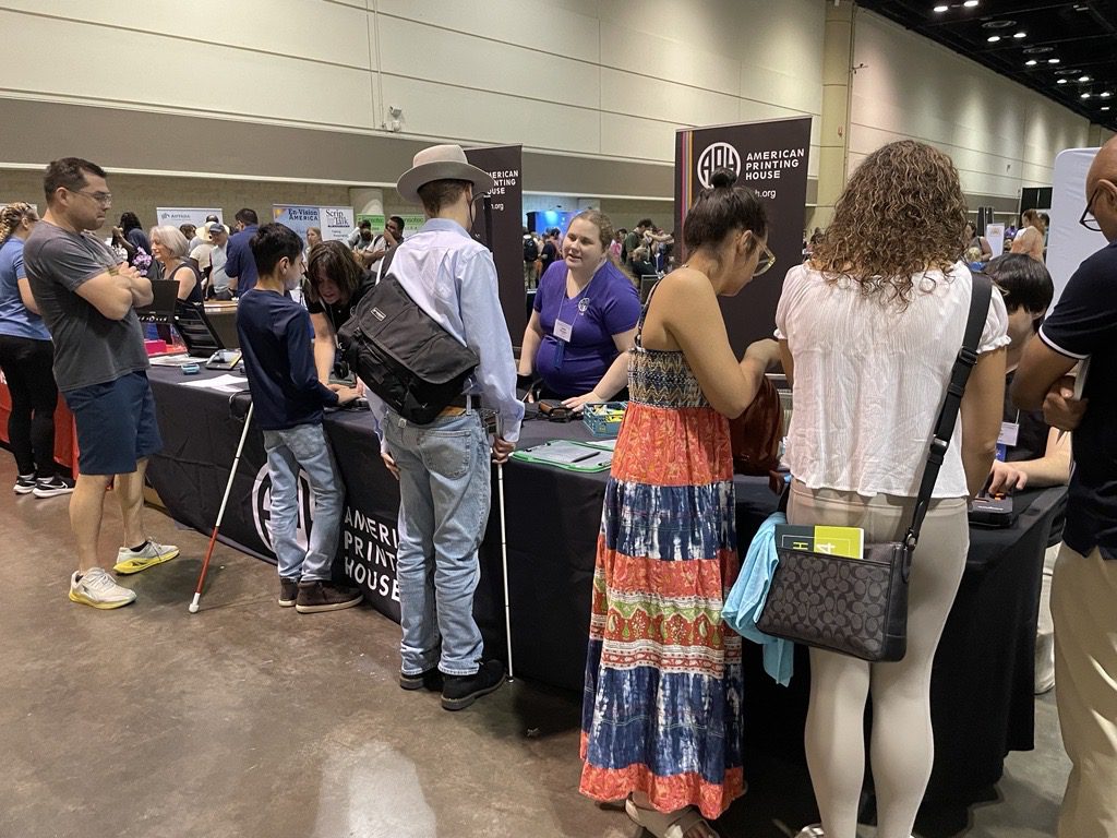 Three APH employees, standing and seated behind a table, speak to several groups of people standing in front of the table.