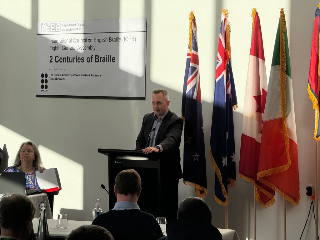An APH employee speaks from behind a podium as a dramatic shadow, likely of a windowpane, can be seen on the wall behind him. To his left are five international flags. To his right, there is a white banner with the words