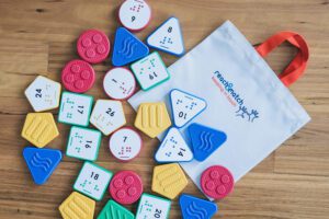 Reach & Match Number Tiles bag filled with multicolored UEB Number Tiles that are spilling out across a wooden surface.