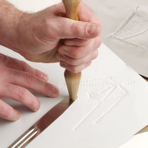 Two hands creating a tactile graphic of shapes and lines using the included hammer and tong tool on white paper.