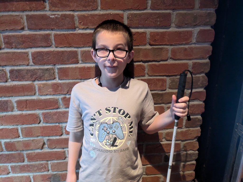 Gavin stands holding his cane and smiling in front of a brick wall.