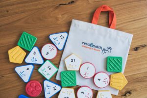 Reach & Match Number Tiles bag filled with multicolored Nemeth Number Tiles that are spilling out across a wooden surface.