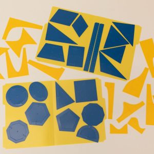 An overhead shot of the Geometric Drawing Stencils attached to a yellow folder with the 3D printed function stencils spread around them. Each shape has an inner stencil and an outer stencil. Shapes include ovals, octagons, circles, triangles, and many more polygons.