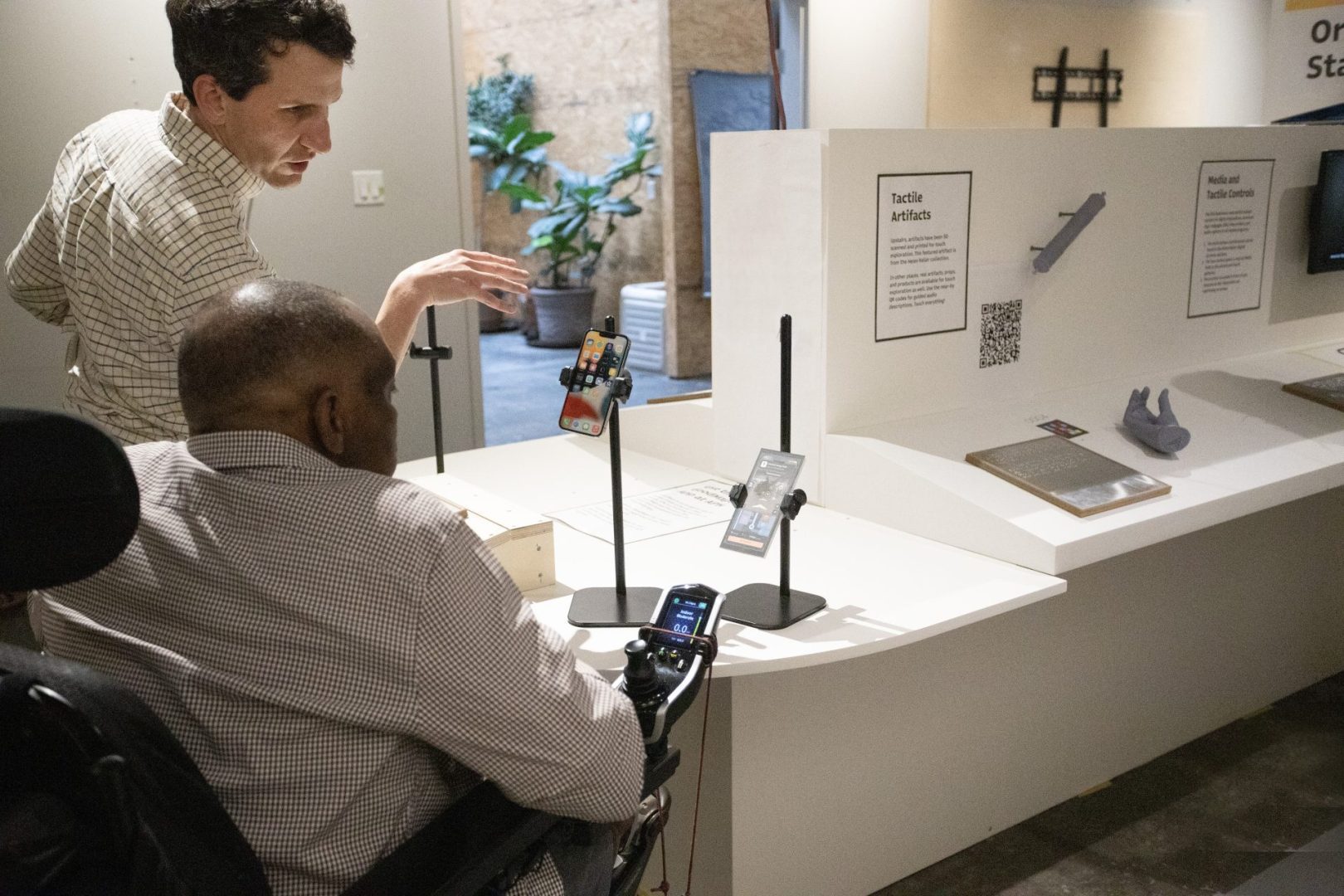 A standing man speaks to a man in a wheelchair while they both face an oval shaped table with a raised section and a short pillar in the middle. In front of the men are two black stands holding smartphones.