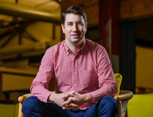Photo of Chris Moizer. He is sitting in a chair with his hands together. He is wearing a red shirt, blue jeans, and brown shoes.