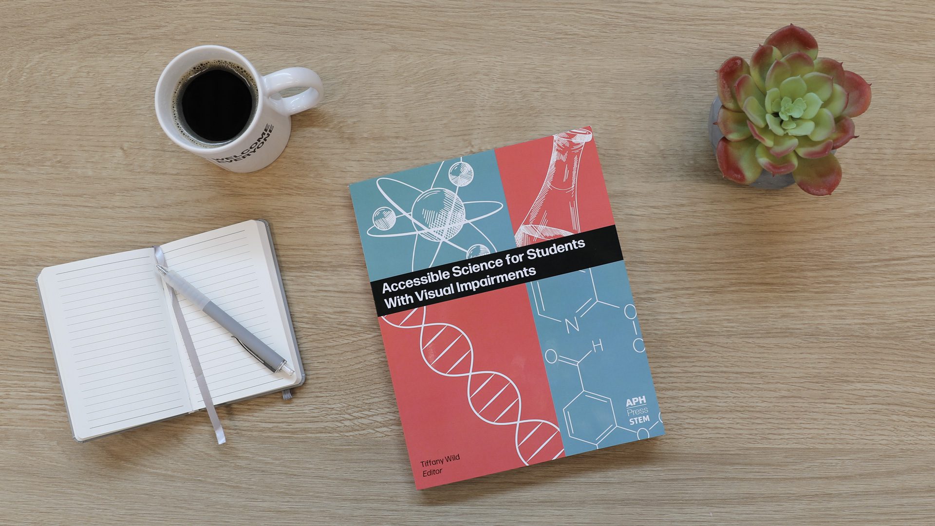A copy of the blue and salmon Accessible Science for Students with Visual Impairments on a wooden table next to a notebook and a mug of black coffee. The cover features white line drawings of scientific diagrams.
