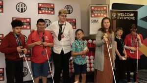 Seven students holding medals and white canes stand in front of a large Abacus and an Abacus Bee step and repeat photo backdrop.