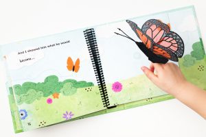 The hand of a young child is playing with a Monarch butterfly manipulative included in On the Way to Literacy: The Caterpillar. The book is opened to a page with bright and colorful illustrations of a butterfly flying over a field.