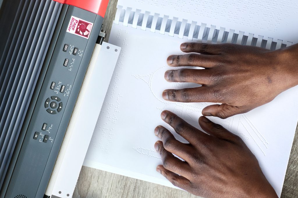 Two hands explore a tactile graphic as it prints from an embosser.
