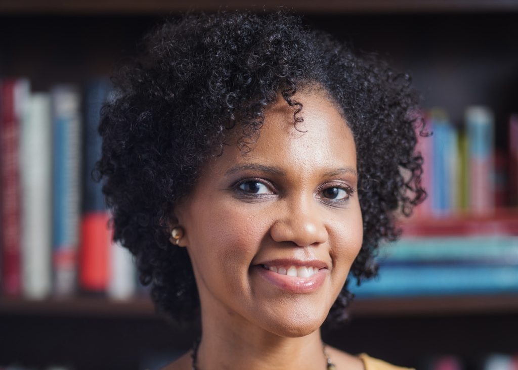 Monique smiles at the camera standing in front of a shelf of books.