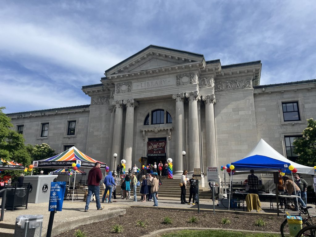 A crowd of people gathers outside of a large concrete pillared building with the words 'Library' etched in stone above the doorway. Colored tents are set up out front and a banner hangs in the doorway with the words 'How-To Festival' in large print on it.