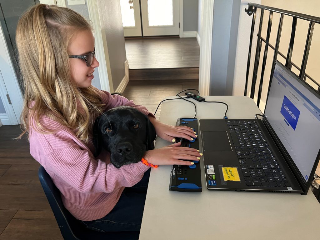 A young girl wearing glasses uses a braille display to read while her dog lays its head on her arm.