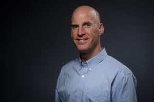 Chris, wearing a blue button down shirt, smiles at the camera.
