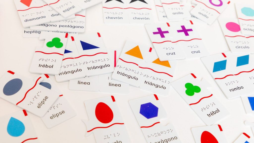 Puzzle cards of various shapes and colors are laid across a table, all with Spanish print and braille labels.