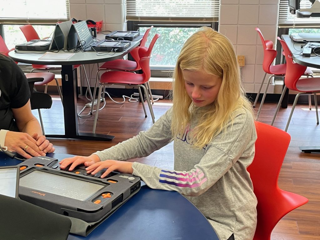 A young girl sits at a classroom desk, with her arms stretched out as she uses her Monarch.