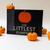 The Littlest Pumpkin storybook stood up on its long edge showing its cover with the title and a simple illustration of an orange pumpkin. Small decorative pumpkins are positioned around and on top of the book.