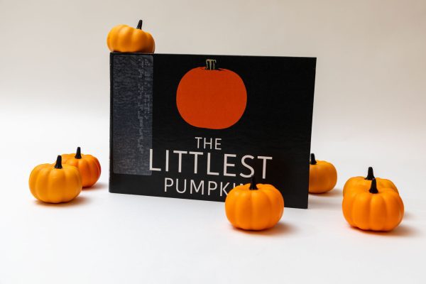 The Littlest Pumpkin storybook stood up on its long edge showing its cover with the title and a simple illustration of an orange pumpkin. Small decorative pumpkins are positioned around and on top of the book.