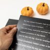 A man's hand holding the left edge of an open page with white large print text and UEB braille. Above the book are two small decorative pumpkins.