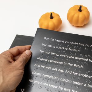 A man's hand holding the left edge of an open page with white large print text and UEB braille. Above the book are two small decorative pumpkins.