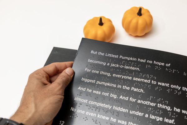 A man's hand holding the left edge of an open page with white large print text and UEB braille. Above the book are two small decorative pumpkins.