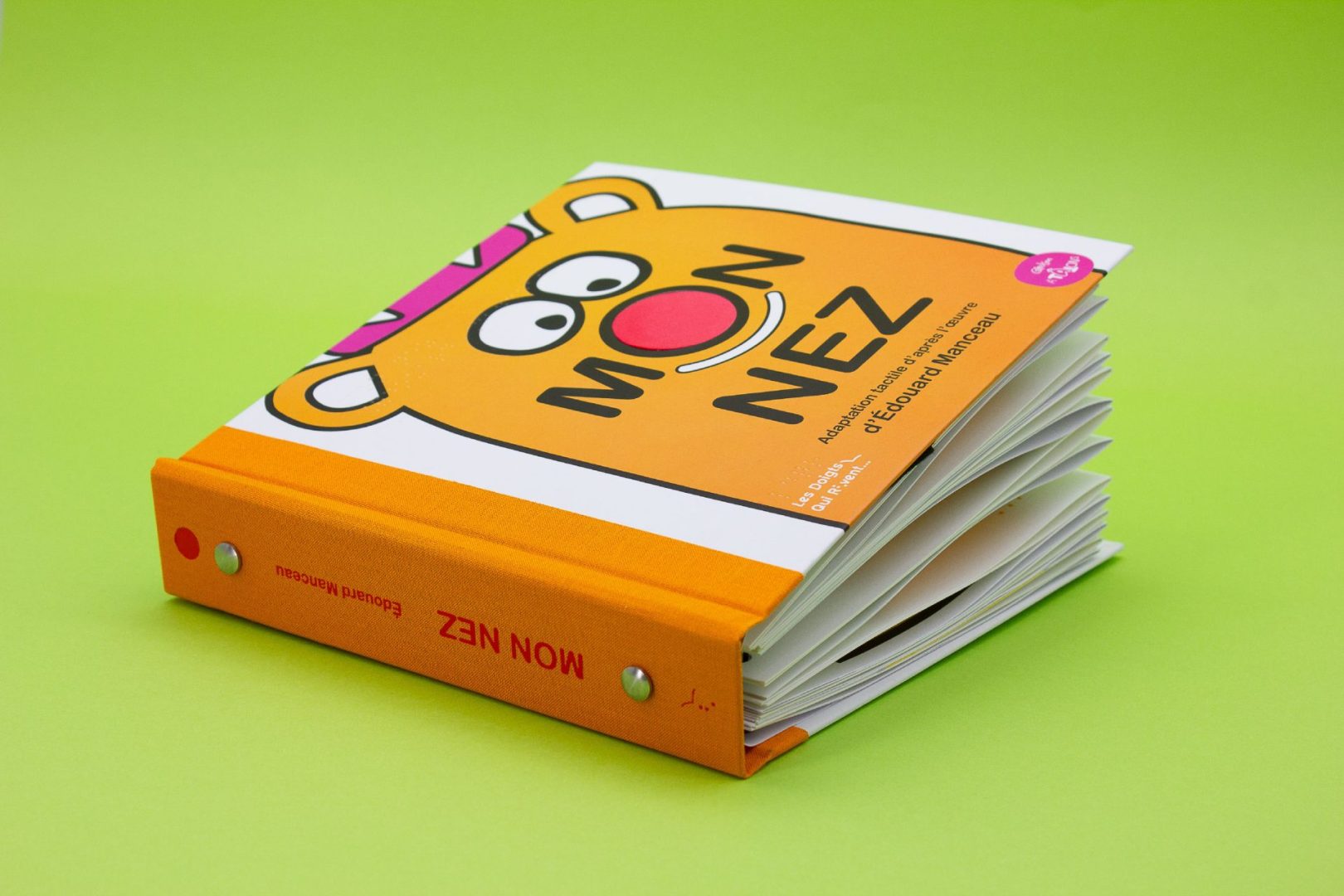 A bright orange book lays against a green background. The book is titled "Mon Nez" but features a big red nose in place of the O. Above the letters are too cartoon eyes, making it look like a face on the cover.