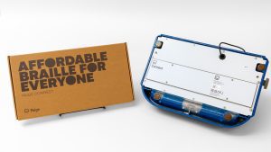 On the right side of the image is a blue Perkins light touch braille writer turned upside down to show the Paige Connect board connected to the base. On the left is the Paige Connect cardboard box that says 
