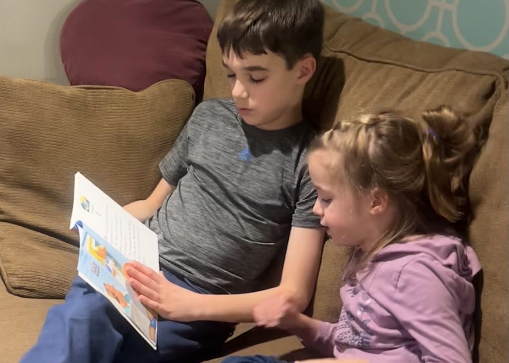 Two students, a boy and a girl sit ont he couch together reading the braille book.