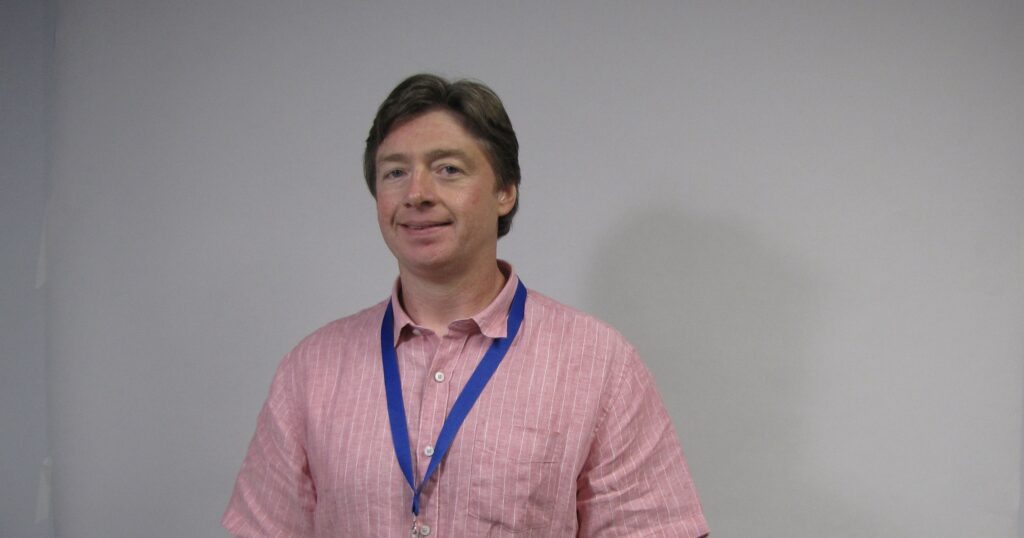 Kris, a man with brown hair, a pink shirt, and a blue APH lanyard, stands smiling in front of a white backgroud.
