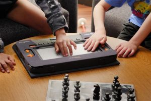 Two students reach out to feel the Monarch screen. Chess pieces rest on the same table in the corner.