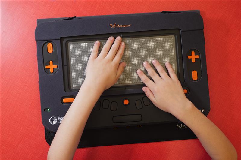A Monarch on top of a red classroom table while two student's hands explore the braille displayed on screen.