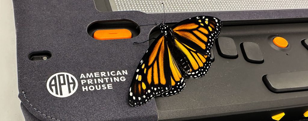 A Monarch butterfly rests on the corner of the Moanrch device, next to the APH logo.